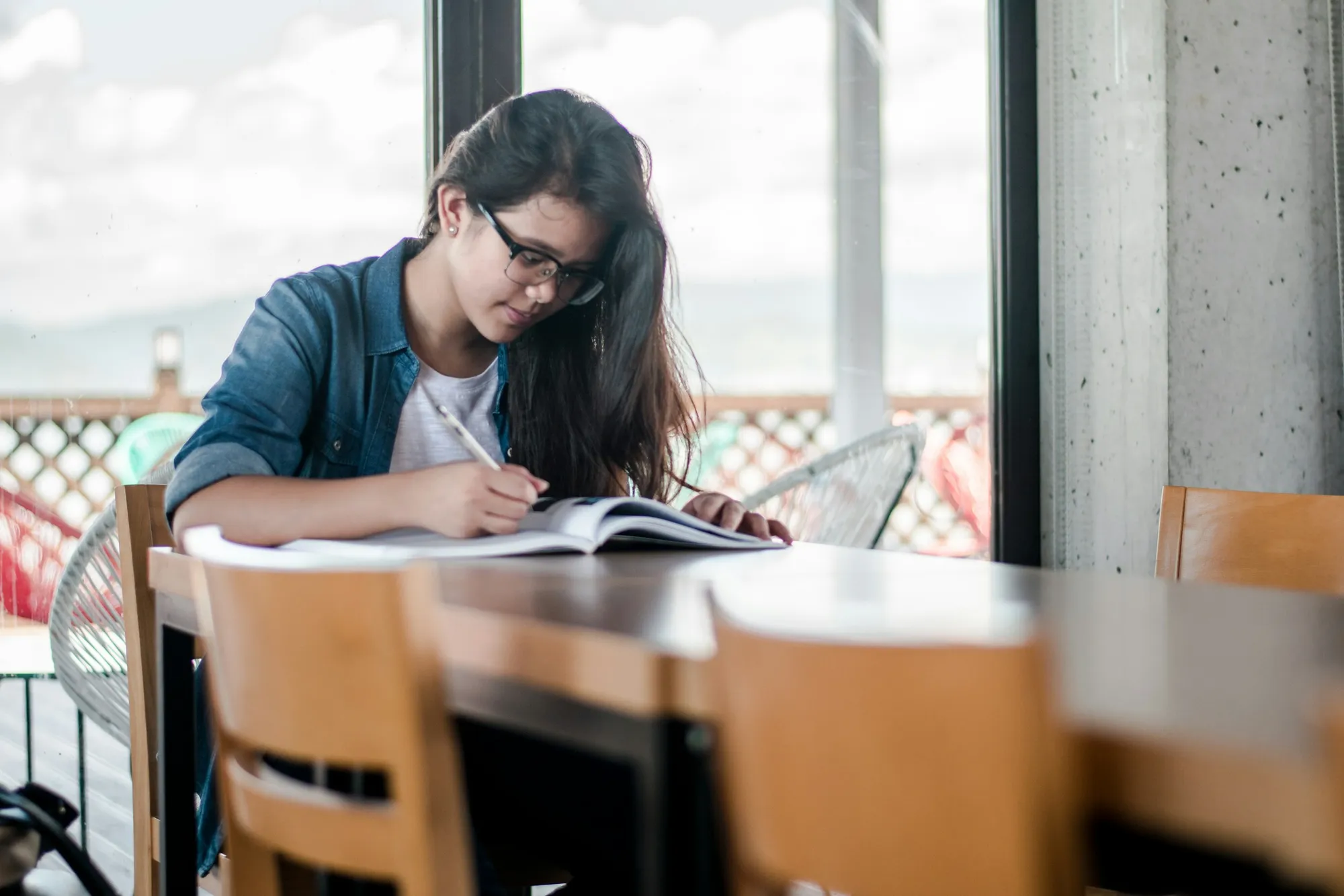 Jovem de óculos e camisa jeans estuda sentada a uma mesa de madeira, perto de uma janela, escrevendo ao lado de um livro aberto