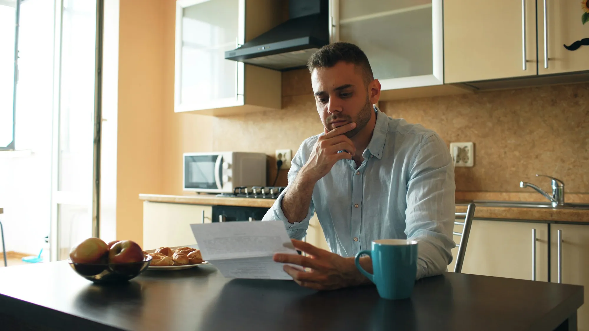 Homem sentado à mesa da cozinha lê uma folha de papel com a mão no queixo, ao lado de uma caneca azul e de uma fruteira com maçãs
