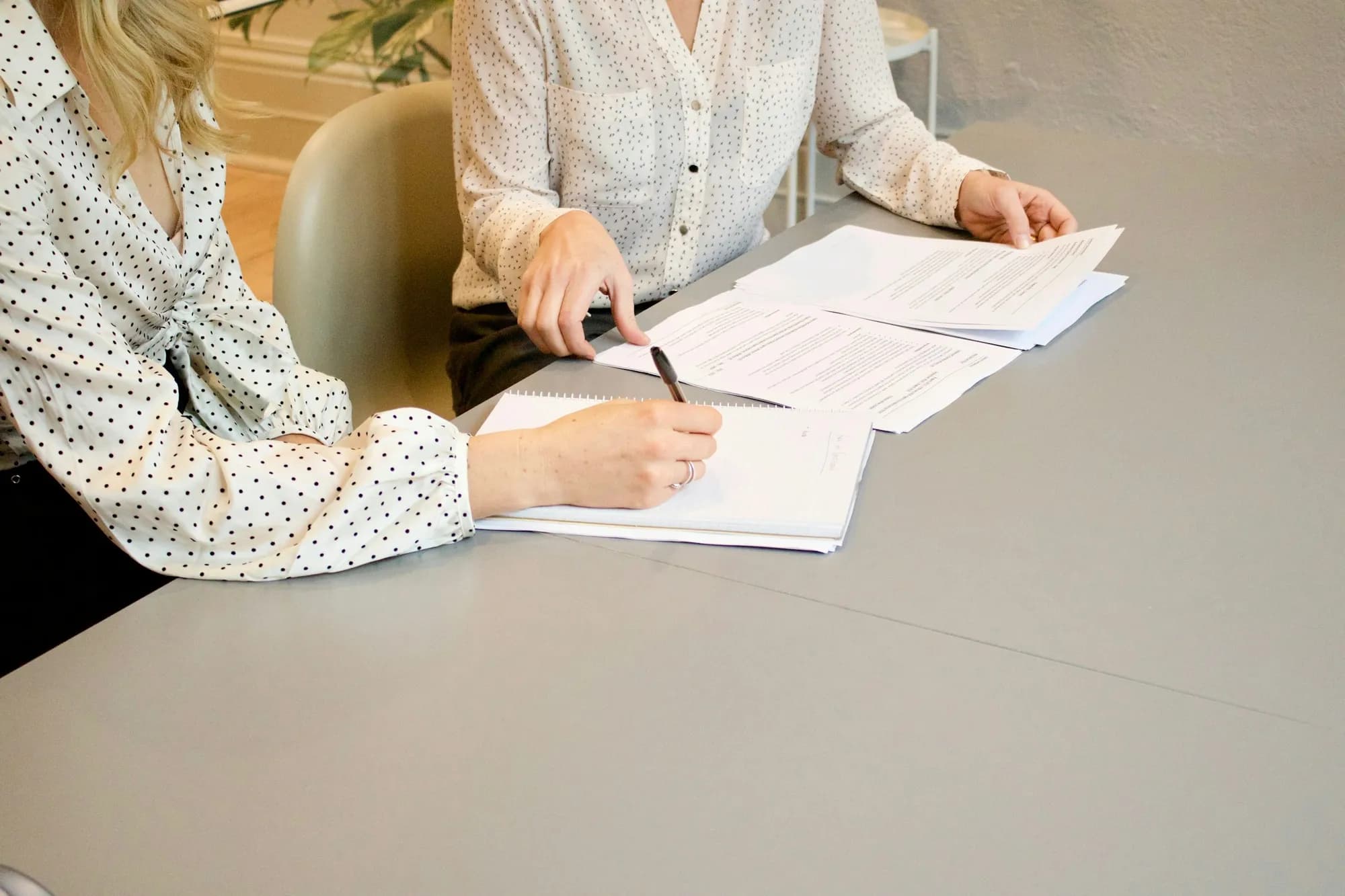 Mulher escreve num caderno sobre uma mesa cinza enquanto outra, ao lado, aponta um trecho de folhas impressas