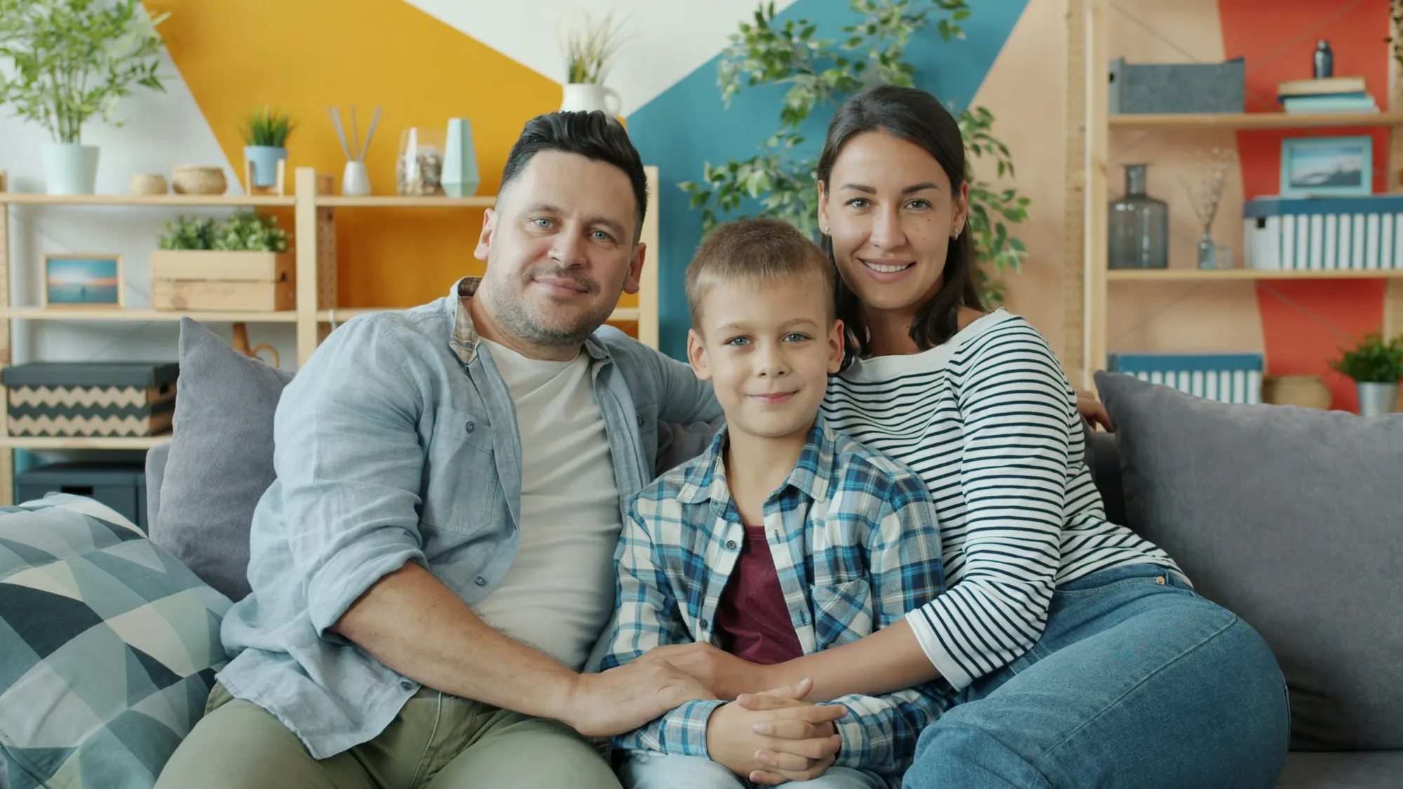 Pai, mãe e filho sentados lado a lado num sofá da sala, sorrindo para a câmera
