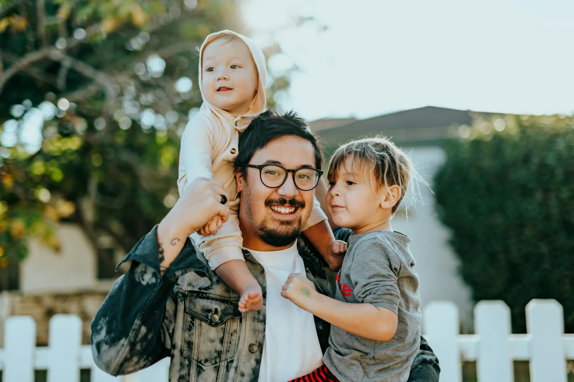 Homem de óculos sorri com uma criança pequena nos ombros e um menino no colo, diante de uma cerca branca